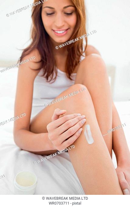 Happy woman applying moisturizer on her leg while sitting on her bed