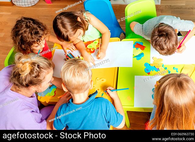 Art lesson at the kindergarten, top view. Lovely children glueing and cutting paper on colorful plastic tables. Children development concept
