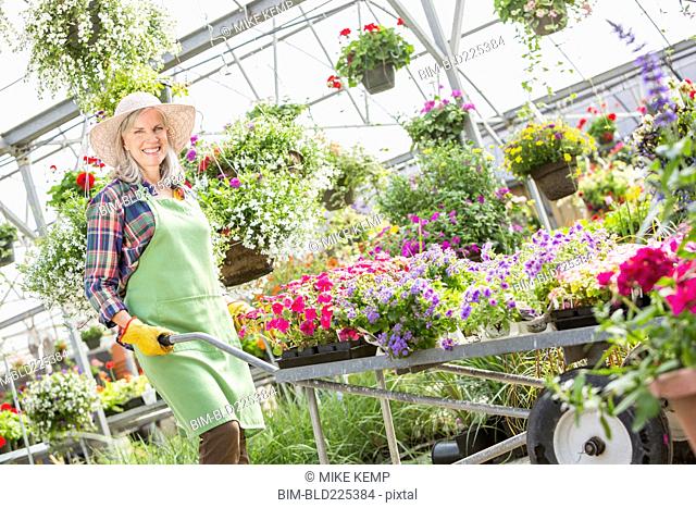 Caucasian woman pushing cart of potted plants in greenhouse