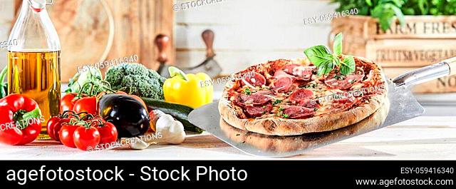 Fresh flame grilled Italian pepperoni or salami pizza served on a metal paddle in a rustic kitchen with an assortment of ingredients in panorama banner format