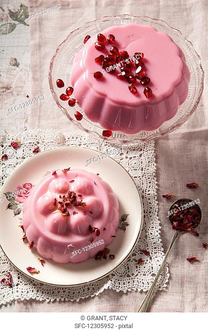 Two pastel pink milk jellies on vintage glass cake stand and plate one covered with pomegrante seeds the other with dried rose petals shot overhead on vintage...