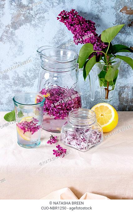 Glass jar of lilac flowers in sugar, glass and pitcher of lilac water with lemon, and branch of fresh lilac on white linen tablecloth with blue textured wall at...