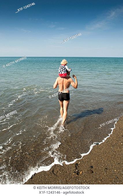 A man carrying his child on shoulders paddling in the sea Aberystwyth wales UK