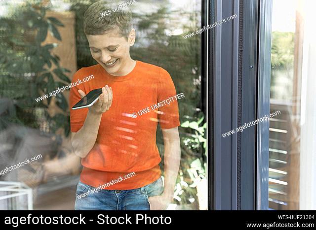 Smiling woman talking over mobile phone while standing by window at home seen through glass