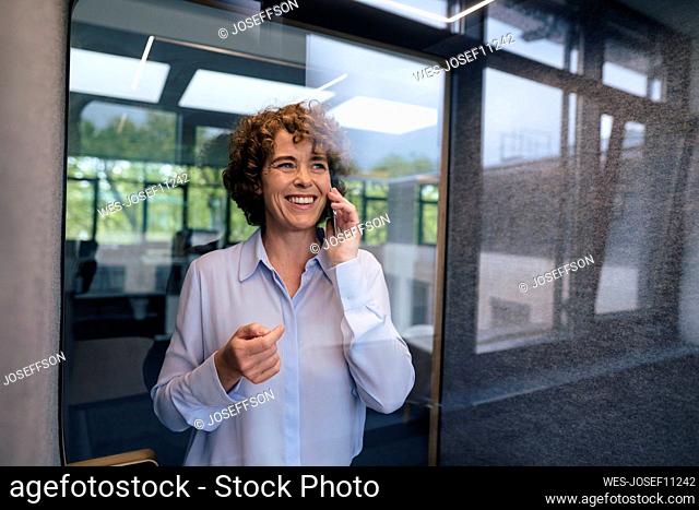 Happy businesswoman talking on smart phone in office seen through glass