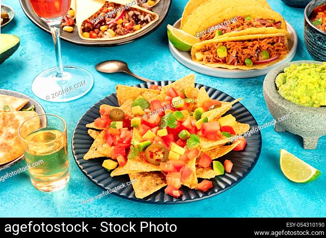 Mexican food with nachos in front and taco shells with pulled pork, burritos, guacamole and other dishes, with tequila and lime on a blue background