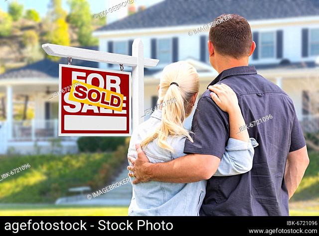 Young adult couple facing front of sold real estate sign and house