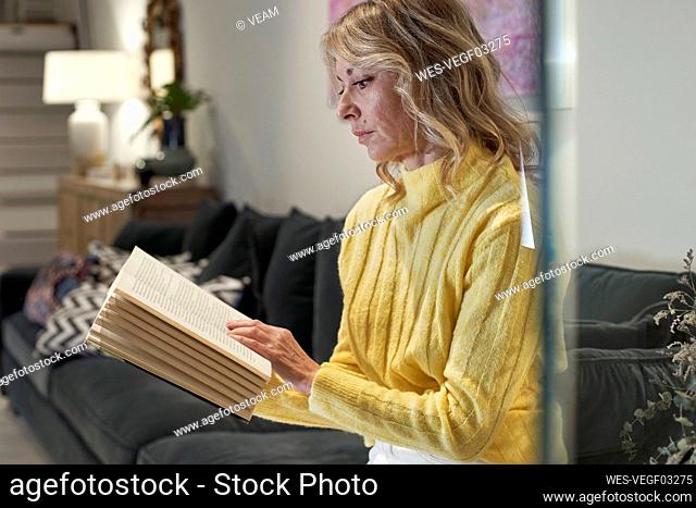 Mature woman in yellow sweater reading book on sofa at home