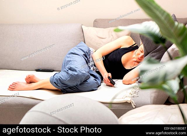 Tired young woman, resting, lying on a comfortable sofa with big cushions in home living room using her mobile phone