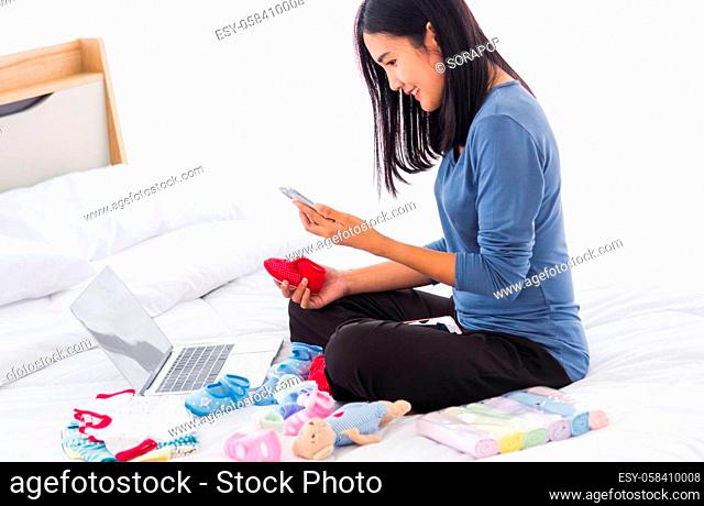 Asian mother shopping online for her baby clothes on laptop computer and tablet on the bed she makes purchase new baby clothes for an unborn baby preparing for...