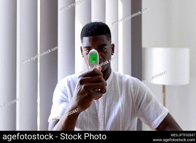 Young man showing temperature on infrared thermometer at home