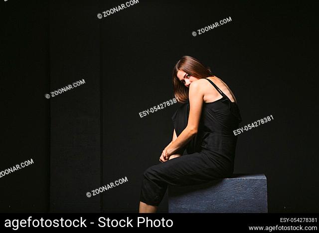 Social theme. young Caucasian woman in strange pose in dress posing on black cube, dark background, symbolizes pain, suffering, seeking help