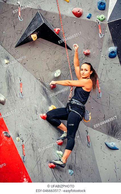 Beautiful young woman in black outfit looking at camera climbing on practical wall in gym, bouldering, extreme sport, rock-climbing concept