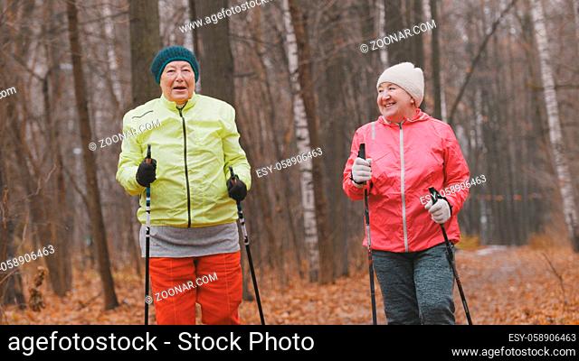 Two elderly woman in autumn park have modern healthy training - nordic walking, telephoto shot