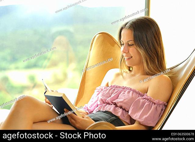 Happy teen reading a paper book sitting on a chair beside a window
