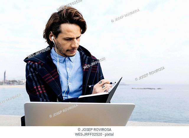 Spain, Barcelona, man sitting at the sea working with laptop and notebook