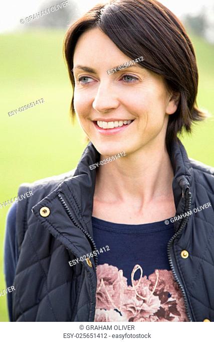 Head and shoulder portrait of a woman looking off into the distance, she is outdoors. A grassy area can be seen behind her, She smiles