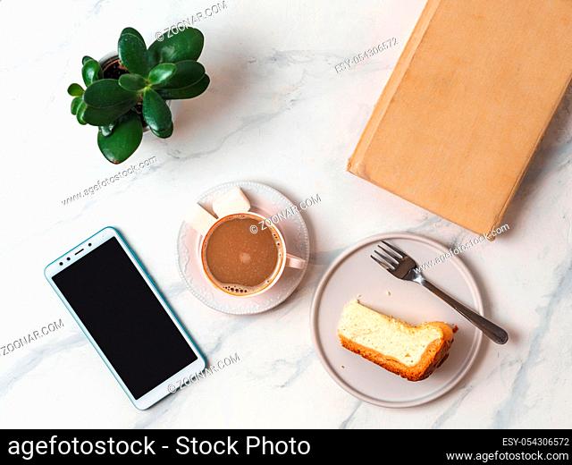 Coffee and cheescake on white marble tabletop. Top view or flat lay. Coffee cup, piece of cheesecake, smartphone, book and succulent