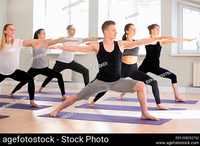 Group of young attractive sporty people practicing yoga lesson, doing Warrior Two exercise, Virabhadrasana II pose, working out indoor full length