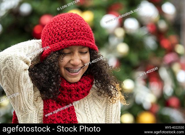 A mixed-race teenage girl sips hot cocoa in front of an outdoor Christmas tree decorated with colorful ornaments. She is weraing hand-knitted hat