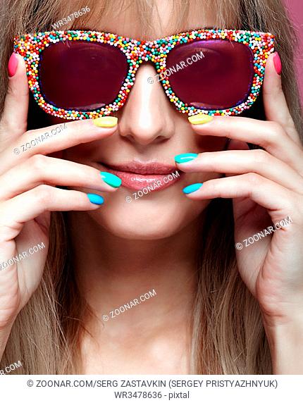Young blonde smiling woman with fun candy glasses and varicoloured finger nails manicure on pink background