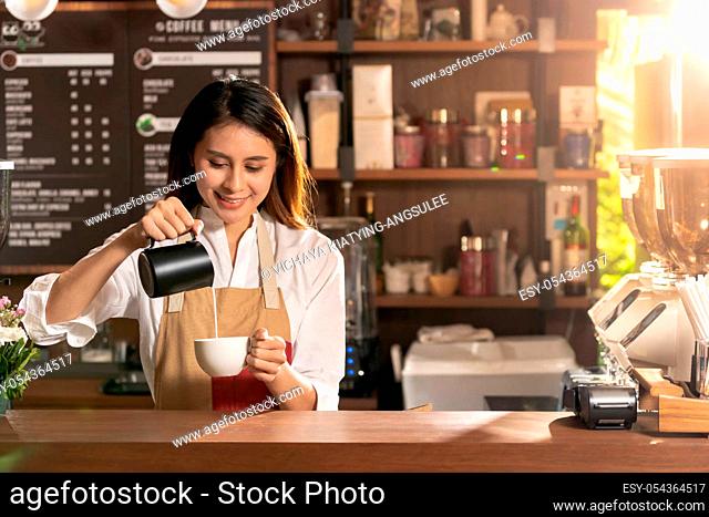 Young adult asian female barista pouring fresh milk to prepare latte coffee for customer in cafe bar