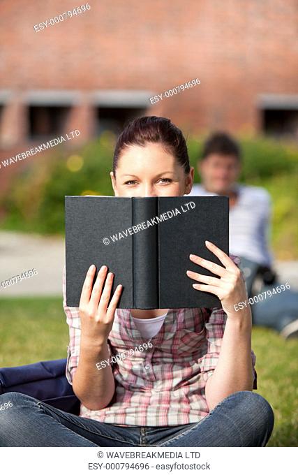 Concentrated female student reading a book sitting on grass in the campus of her university
