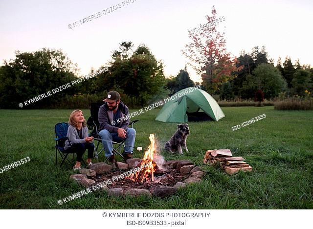 Father and daughter sitting beside campfire, young girl toasting marshmallows over fire