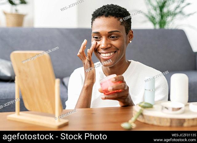 Smiling woman using facial cream and looking in mirror at home