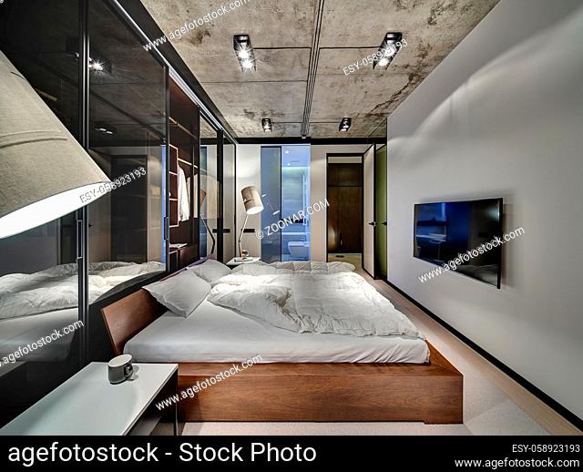 Room in a loft style with white walls and concrete ceiling. There is a TV, bed with pillows and blankets, glowing lamps, wardrobe with glass sliding doors