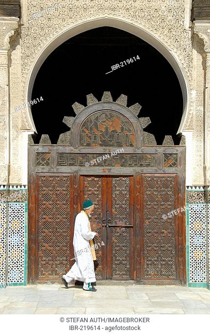 Man wearing traditional cloth in front of an old carved wooden gate Medersa Bou Inania Fes El-Bali Morocco