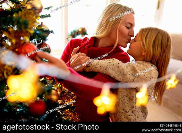 Front view of a young Caucasian woman kissing her young daughter beside the Christmas tree in their sitting room, with Christmas star decorations hanging in the...