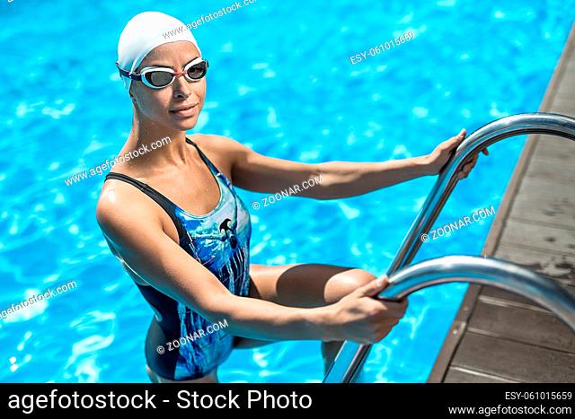 Smiling sportive girl stands on the pool ladder in the swim pool and holds hands on it. She wears a blue-black swimsuit, a white swim cap and swim glasses