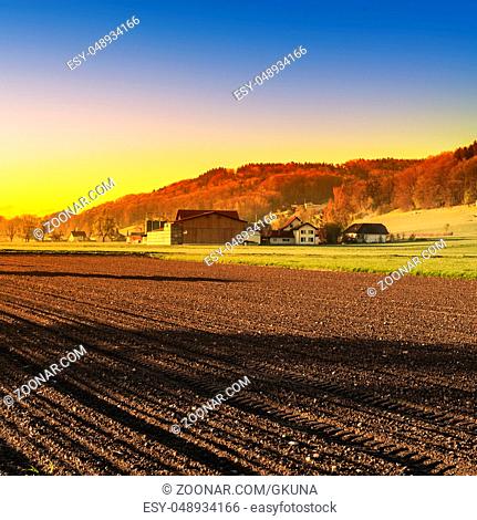 Swiss village surrounded by forests and plowed fields at sunset. Agriculture in Switzerland, arable land and pastures