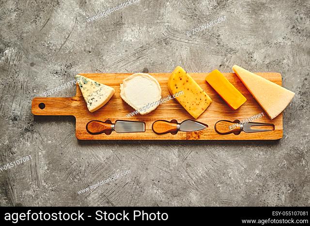Assortment of various kinds of cheeses served on wooden board with fork and knives. Placed on concrete background