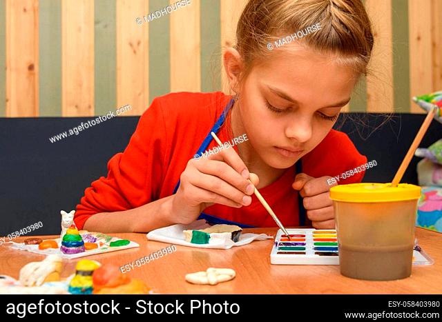 A girl dips a brush in paint of the desired color while painting a craft from salt dough