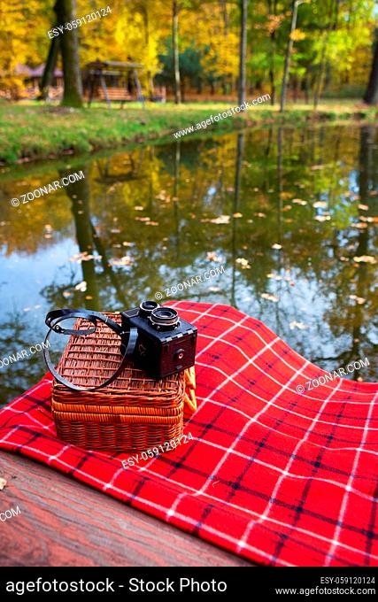 The old camera lies on a suitcase on a red plaid blanket. Wooden bridge near the lake