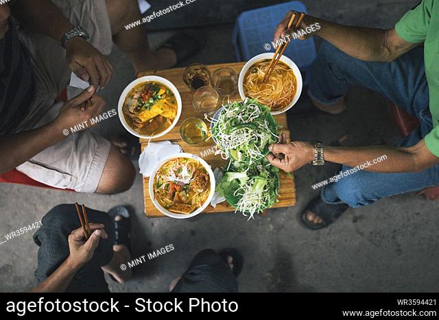 High angle close up of three men tucking into bowls of noodles for breakfast in a small alleyway