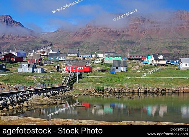 Simple bridge leading to houses reflected in a body of water, dissipating fog and red volcanic rock, wall of fog, red volcanic rock, Disko Island, Disko Bay