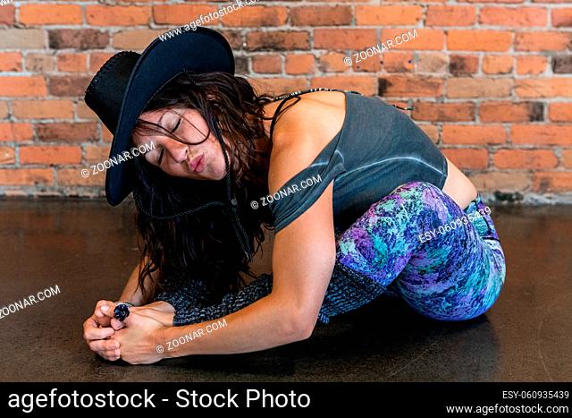 A close up shot of a fun and flexible lady in her 30s, in a seated yoga pose as she blows a kiss with pursed lips, wearing large brimmed hat