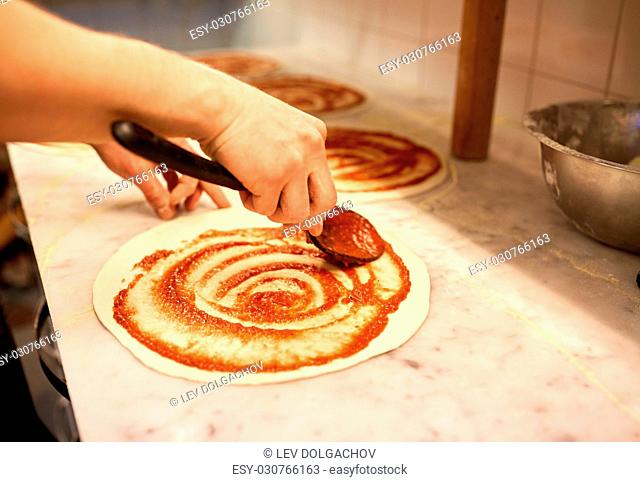 food, culinary, italian cuisine, people and cooking concept - cook with spoon applying tomato sauce to raw pizza dough at pizzeria