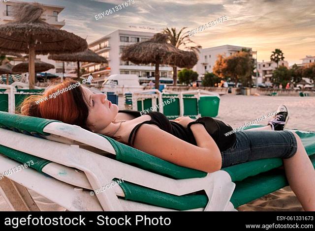 Young woman with long red and black colored hair lying down on a deckchair at the beach, looking at camera, , side view, horizontal