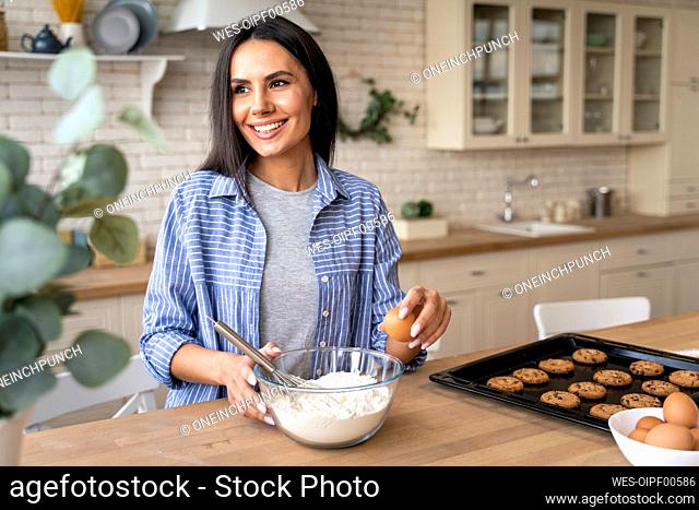 Smiling woman looking away holding egg while preparing food in kitchen at home