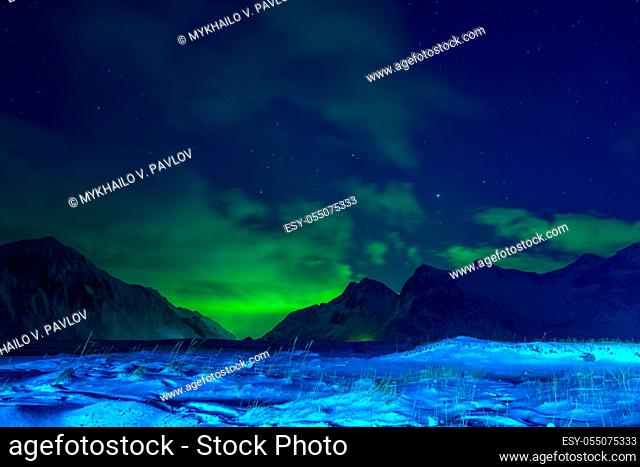 Norway. Lofoten. Winter night. Snow-covered grass and mountains. Clouds in the sky and northern lights