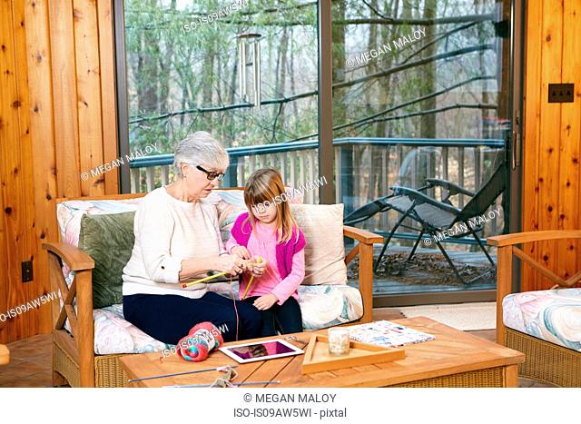 Senior woman teaching granddaughter how to knit on living room sofa