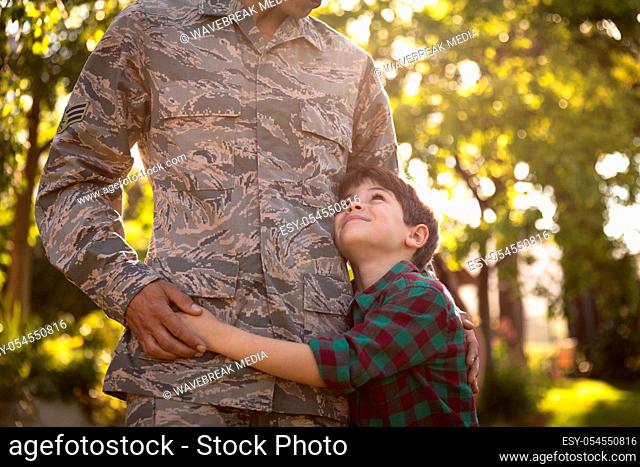 Front view mid section of a young adult mixed race male soldier in the garden outside his home, embracing his young son, who is looking up at him smiling