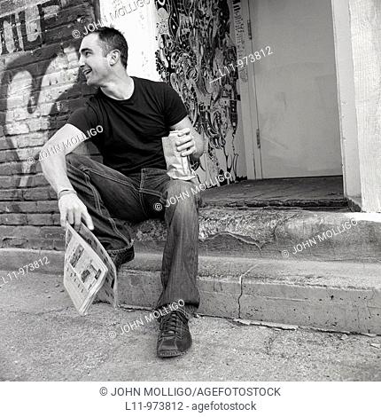 Man seated in alley, with newspaper and beer