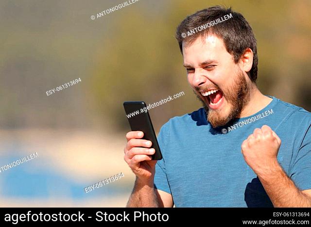 Excited man checking good news on mobile phone outdoors in nature