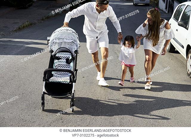 parents with daughter on hands, walking on street, with buggy, parenthood, family, one child, candid, unposed. In holiday destination Chersonissos, Crete