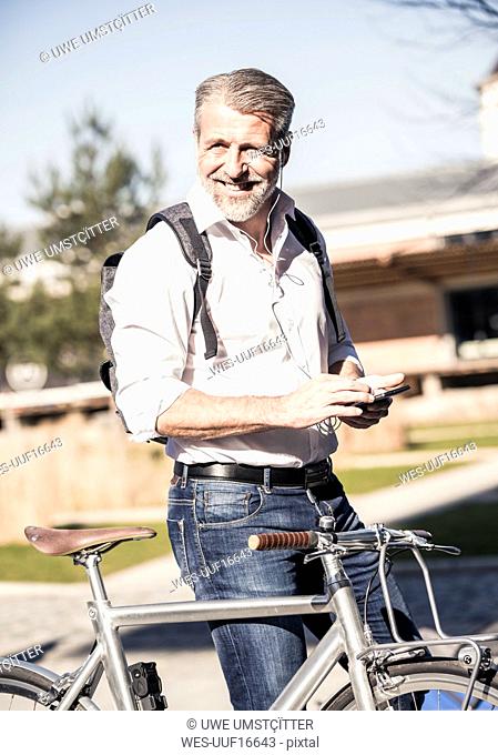 Portrait of smiling mature businessman with bicycle, cell phone and earphones